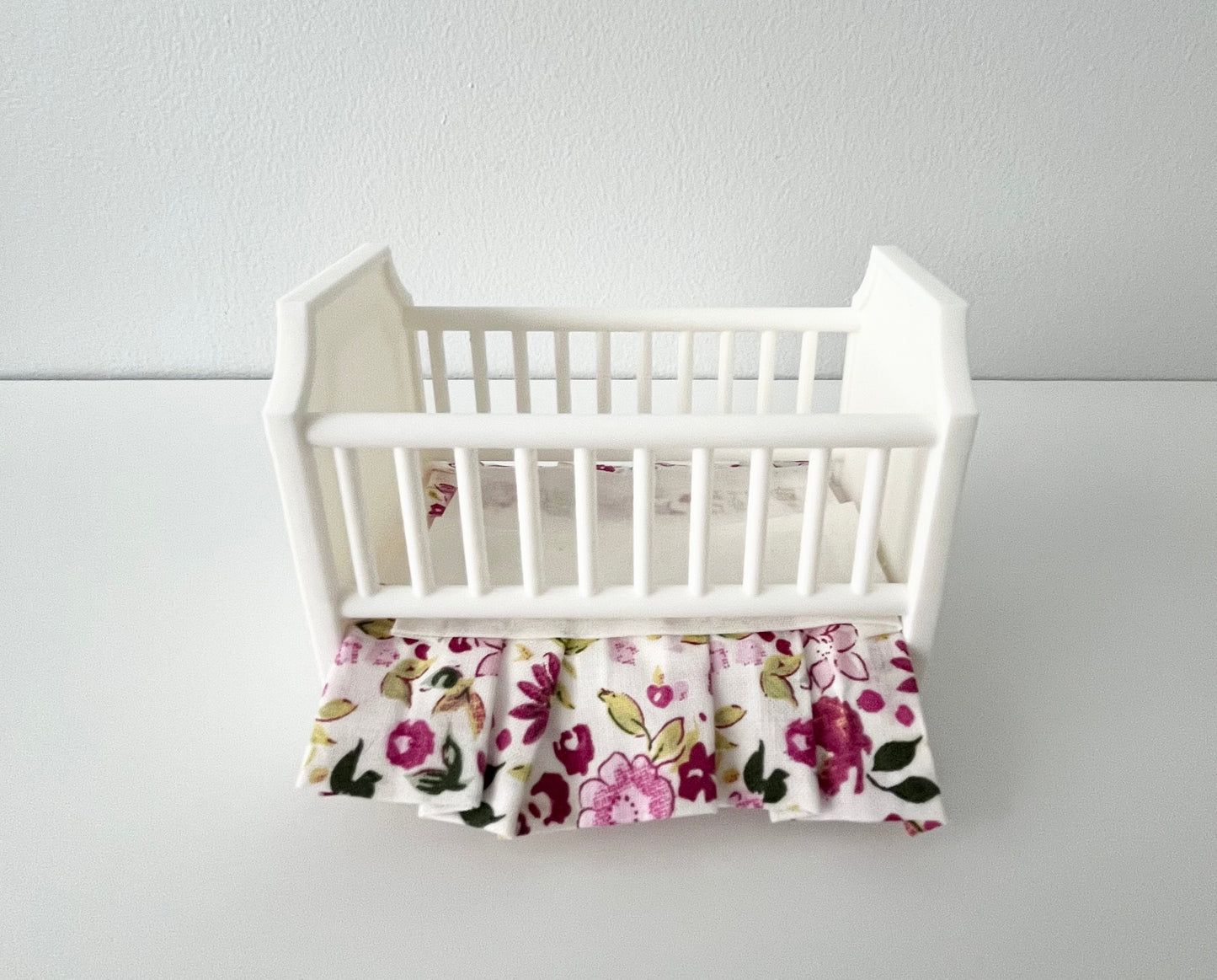 Front view of White baby crib in 1:12 scale with floral bed skirt on a light gray background